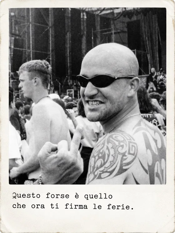 Un uomo con la testa rasata, occhiali da sole e la spalla tatuata sorride davanti al palco e alza il dito medio.